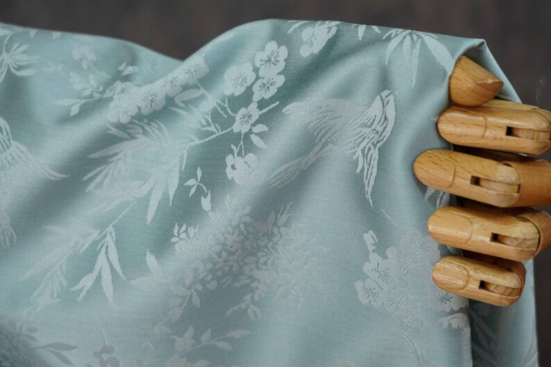 May include: Light blue fabric with a white floral and bird pattern. The fabric is draped, showing the intricate design of flowers, leaves, and birds. The fabric is held by a wooden hand.