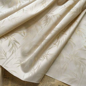 May include: Cream-coloured fabric featuring a delicate bamboo leaf design. The fabric is folded, revealing its texture and the interplay of light and shadow. The leaves are a pale gold, offering a refined aesthetic. Suitable for interior design.