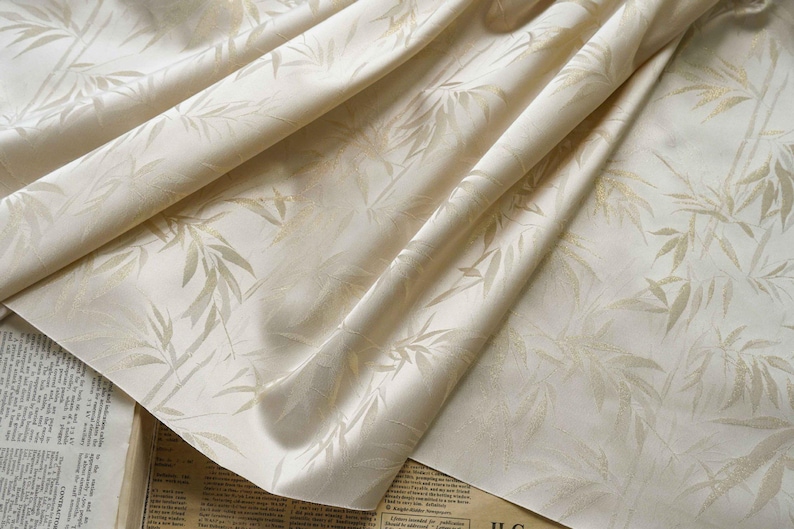 May include: Cream-coloured fabric featuring a delicate bamboo leaf design. The fabric is folded, revealing its texture and the interplay of light and shadow. The leaves are a pale gold, offering a refined aesthetic. Suitable for interior design.