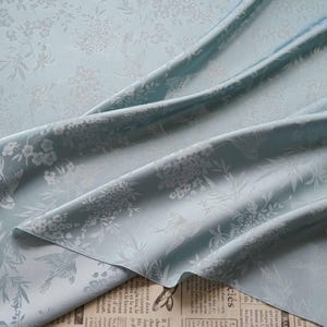 May include: A close-up of a light blue fabric with a floral and bird pattern. The fabric is draped, showing folds and texture. The design features white flowers and birds, creating a delicate and elegant aesthetic.