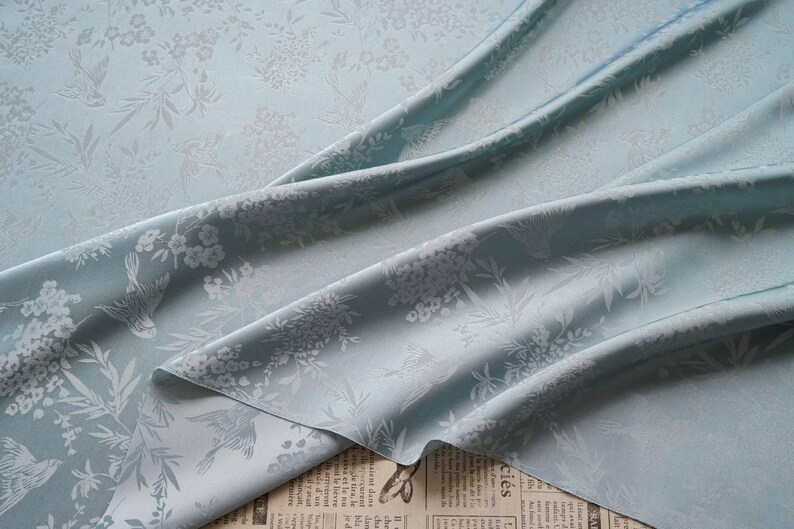 May include: A close-up of a light blue fabric with a floral and bird pattern. The fabric is draped, showing folds and texture. The design features white flowers and birds, creating a delicate and elegant aesthetic.