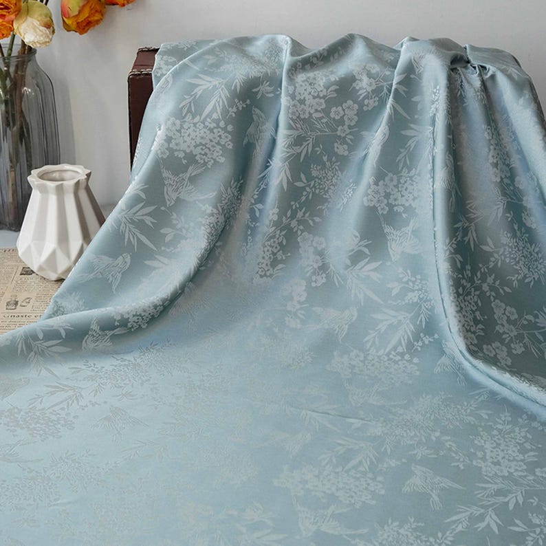 May include: A light blue fabric with a floral and bird pattern. The fabric is draped, showing the intricate white design of flowers, leaves, and birds. The material has a subtle sheen, suggesting a luxurious texture.