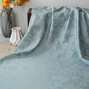 May include: A light blue floral patterned fabric draped over a table. The fabric has a subtle sheen and a delicate floral design. A vase of orange flowers and a decorative vase are in the background.