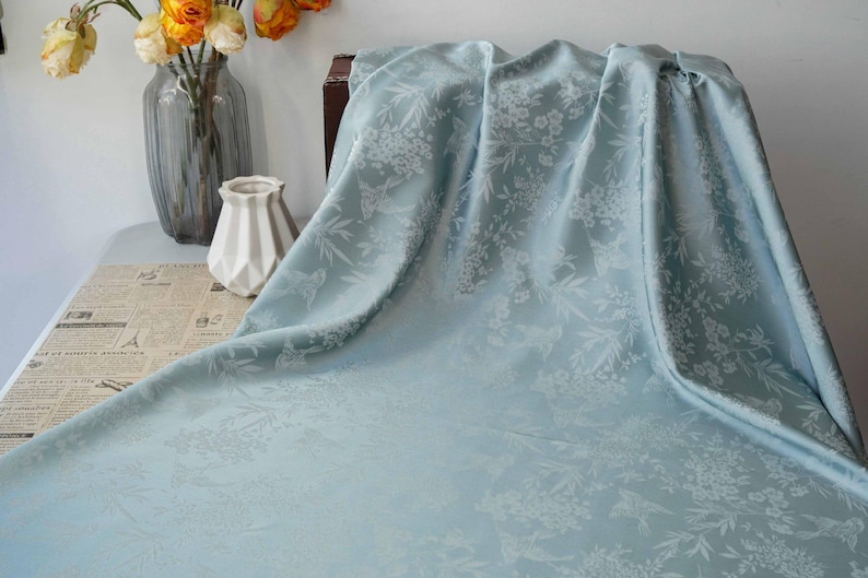 May include: A light blue floral patterned fabric draped over a table. The fabric has a subtle sheen and a delicate floral design. A vase of orange flowers and a decorative vase are in the background.