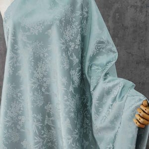 May include: A light blue fabric with a floral and bird pattern. The fabric is draped over a mannequin, showcasing its texture and design. The pattern features white flowers and birds, creating a delicate and elegant aesthetic.
