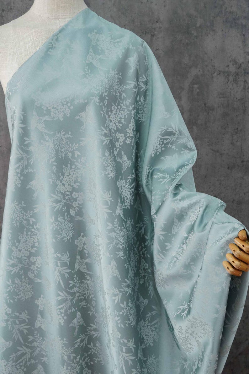 May include: A light blue fabric with a floral and bird pattern. The fabric is draped over a mannequin, showcasing its texture and design. The pattern features white flowers and birds, creating a delicate and elegant aesthetic.