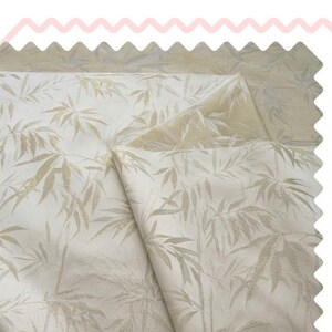 May include: Cream-coloured fabric featuring a repeating pattern of bamboo leaves in a pale gold hue. The fabric is folded, showing a lighter gold backing. The edges are scalloped, and the overall look is soft and textured.