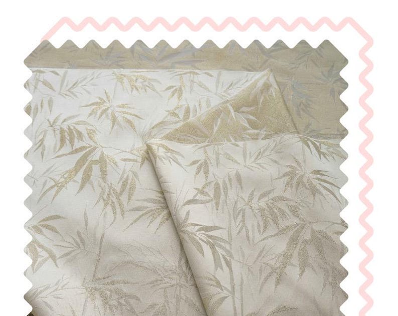May include: Cream-coloured fabric featuring a repeating pattern of bamboo leaves in a pale gold hue. The fabric is folded, showing a lighter gold backing. The edges are scalloped, and the overall look is soft and textured.