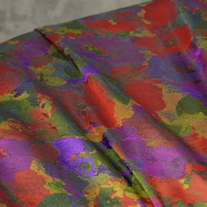 May include: A piece of fabric featuring a vibrant, abstract floral pattern with overlapping patches of deep red, bright orange, vivid purple, and olive green. The material has a subtle sheen and a textured weave, creating a complex, multi-coloured surface.
