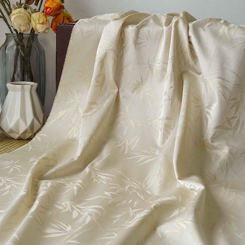 May include: Cream-coloured fabric featuring a delicate bamboo leaf design. The fabric is draped, forming gentle folds and shadows. The texture appears smooth and luxurious, ideal for home decor or clothing.