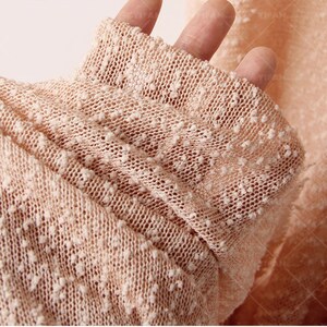 May include: A light peach-colored knit fabric featuring a textured, nubby surface with small, raised white flecks throughout the material. The textile has a semi-sheer, open-weave construction that creates a soft, dimensional appearance across the surface.