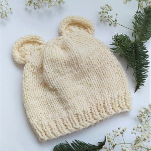 May include: Cream-colored knitted hat with two small, rounded ear-like protrusions. The hat is made of a thick, textured yarn and has a ribbed brim. The hat is likely for a baby or child. The background includes sprigs of white flowers and a green pine branch.