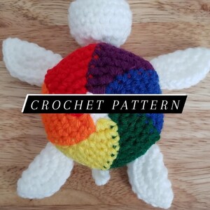 May include: A crocheted rainbow turtle with white wings. The turtle is made with different colored yarn, including red, orange, yellow, green, blue, and purple. The text "CROCHET PATTERN" is visible on the image.