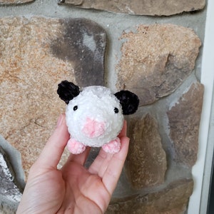 May include: A small white and black crocheted plush toy resembling an opossum. The toy has pink ears, a pink nose, and pink feet.