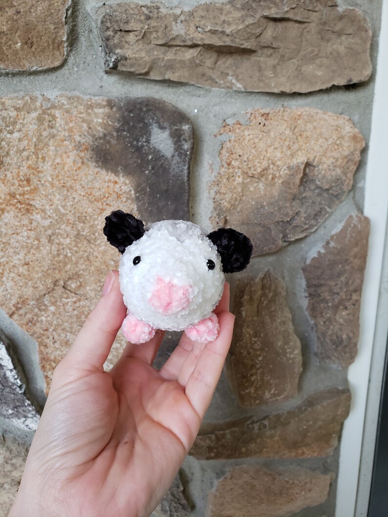 May include: A small white and black crocheted plush toy resembling an opossum. The toy has pink ears, a pink nose, and pink feet.