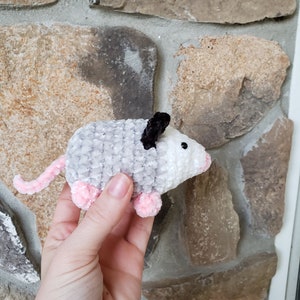 May include: A small, grey and white crocheted plush toy shaped like an opossum. The toy has pink feet and black ears.