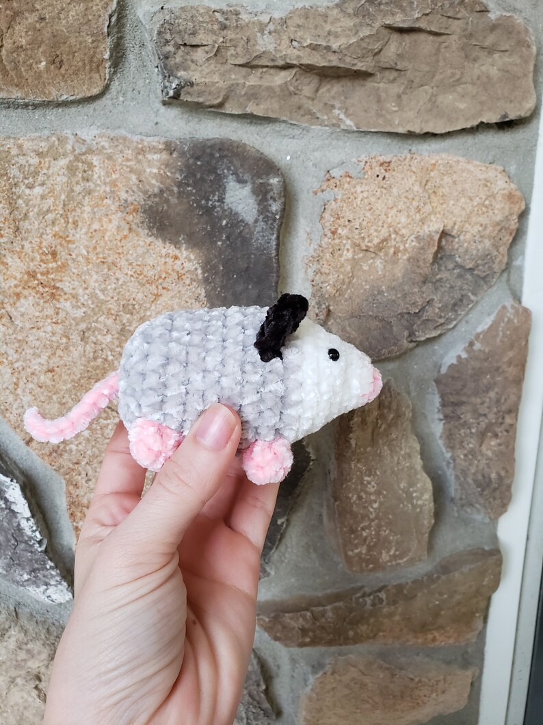 May include: A small, grey and white crocheted plush toy shaped like an opossum. The toy has pink feet and black ears.