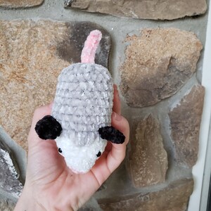 May include: A crocheted plush toy resembling an opossum. The toy is grey and white with black ears and a pink tail. The toy is being held in a person's hand.