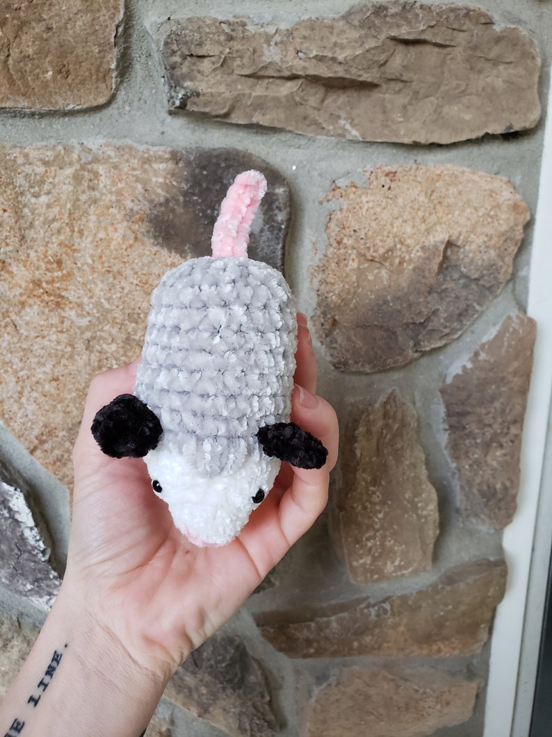 May include: A crocheted plush toy resembling an opossum. The toy is grey and white with black ears and a pink tail. The toy is being held in a person's hand.