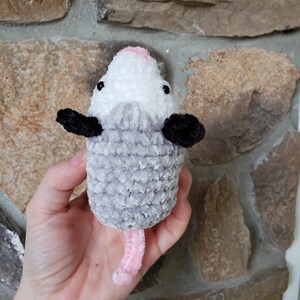 May include: A grey and white crocheted plush toy shaped like an opossum with a pink nose and tail. The toy is being held in a person's hand.