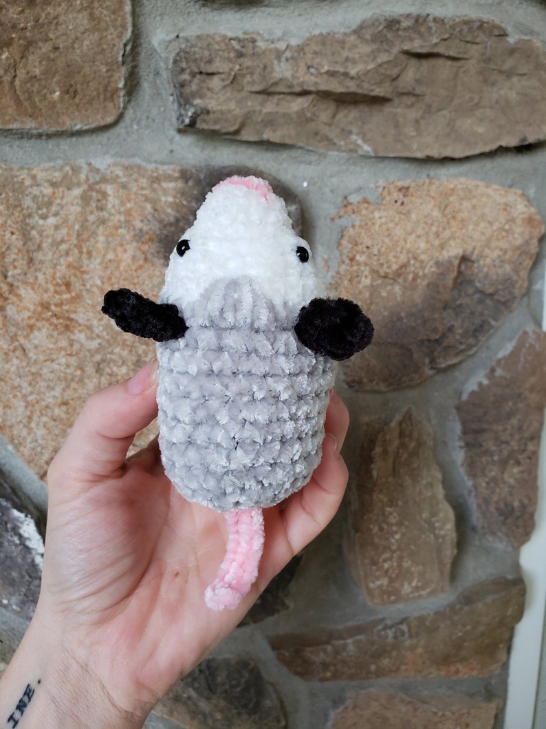 May include: A grey and white crocheted plush toy shaped like an opossum with a pink nose and tail. The toy is being held in a person's hand.