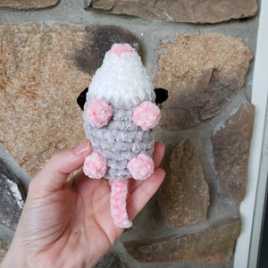 May include: A crocheted plush toy shaped like a possum. The toy is grey, white, and pink with black eyes. The possum is sitting on its hind legs with its tail curled up behind it.
