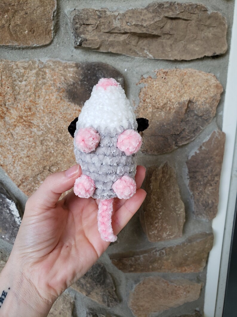 May include: A crocheted plush toy shaped like a possum. The toy is grey, white, and pink with black eyes. The possum is sitting on its hind legs with its tail curled up behind it.