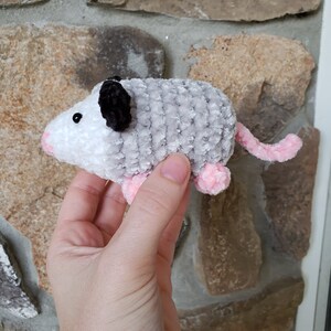May include: A small crocheted plush toy shaped like an opossum. The opossum is grey and white with pink feet and a black nose. It has a black and white crocheted eye.