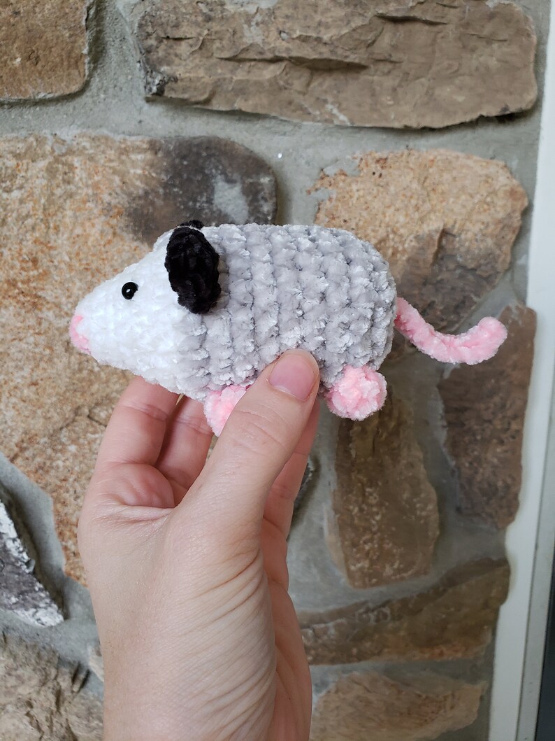 May include: A small crocheted plush toy shaped like an opossum. The opossum is grey and white with pink feet and a black nose. It has a black and white crocheted eye.