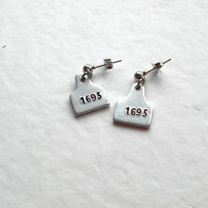May include: A pair of silver earrings shaped like livestock tags with the number 1695 printed on each tag.
