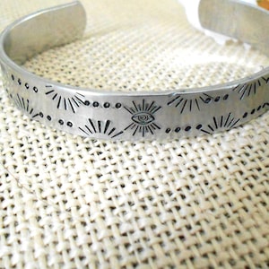 May include: A silver-colored cuff bracelet with an engraved design. The design includes an eye symbol, sunburst patterns, and dotted lines. The bracelet is open-ended and appears to be made of metal.