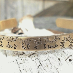 May include: A gold-colored cuff bracelet with the phrase "one day at a time" engraved in a handwritten font. The bracelet is decorated with sun and flower designs.