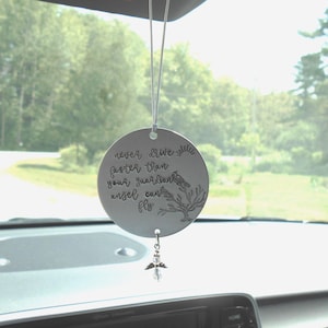 May include: A silver-colored car charm with the engraved text "never drive faster than your guardian angel can fly." The charm features a bird and tree design, with a small angel pendant hanging below.