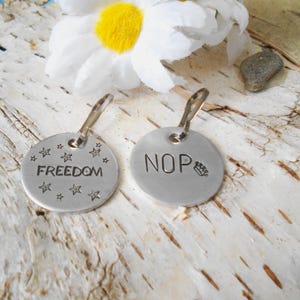 May include: Two silver, round pet tags with attached metal loops. One tag reads "FREEDOM" with star accents. The other tag reads "NOP" with a paw print. The tags are displayed on a textured, light-colored surface with a white flower.