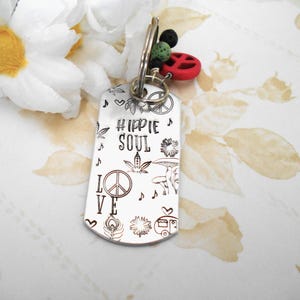 May include: A silver-colored metal keychain tag with the words "Hippie Soul" and the word "Love" etched on it. The tag is decorated with peace signs, musical notes, and other hippie-themed designs. A red peace sign charm and black and green beads are attached.