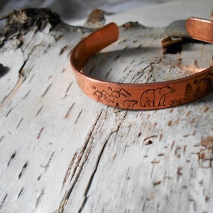 May include: A copper cuff bracelet with an engraved bear and mountain scene. The open-ended bracelet is made of polished copper and features a detailed forest and wildlife design. The bracelet is approximately 2.5 inches in diameter.