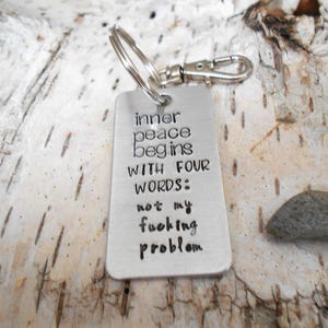 May include: A silver metal keychain tag with the text "inner peace begins with FOUR WORDS: not my fucking problem." The keychain has a silver clasp and ring. The background is a piece of birch bark.