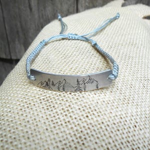 May include: A silver-toned metal bar bracelet featuring a black engraved mountain and pine tree landscape design. The bracelet is attached to a light blue braided cord with an adjustable sliding knot closure for a secure and flexible fit.