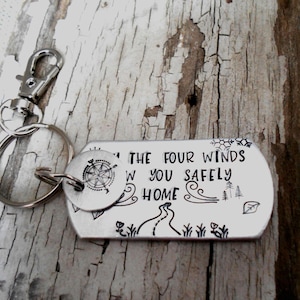 May include: A silver-colored keychain with a rectangular tag and a round tag. The rectangular tag has the text "...THE FOUR WINDS W YOU SAFELY HOME" with a compass design and other illustrations. The keychain is on a wooden surface.