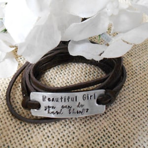 May include: A wrap bracelet with a dark brown leather band and a silver rectangular plate. The plate is stamped with the words "Beautiful Girl you can do hard things". White flowers are in the background.