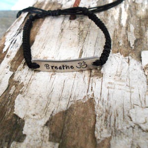 May include: A black cord bracelet with a silver metal bar. The bar is engraved with the word "Breathe" and the Om symbol. The bracelet is displayed on a weathered, light brown wooden surface.