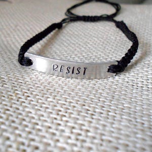 May include: A black braided bracelet with a silver metal tag that says "RESIST".