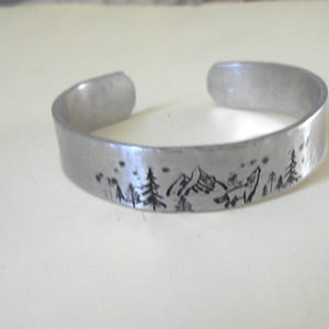 May include: A silver-colored cuff bracelet with a nature-themed design. The bracelet features a mountain range, trees, and a fox silhouette etched into the metal. The bracelet is open-ended and appears to be made of a lightweight metal.