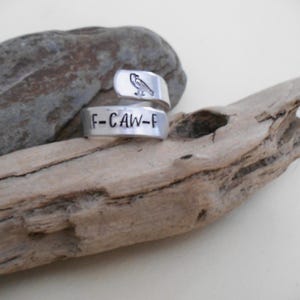 May include: A silver-colored wrap ring with a bird design and the text "F-CAW-F". The ring is displayed on a piece of driftwood. The bird design is on the top band of the ring.