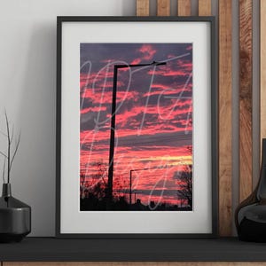 May include: Framed print depicting a sunset with a sky of pink and red clouds. Silhouetted streetlights and trees are set against the colorful sky. The print is in a black frame, displayed on a black shelf.