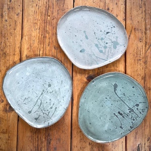 May include: Three ceramic plates with a speckled glaze in shades of grey and blue. The plates are irregularly shaped and have a rustic, handmade look.