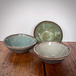 May include: Three ceramic bowls in various shades of green and brown. The bowls are handmade and have a rustic, earthy feel.