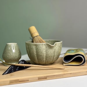 May include: A matcha tea set on a wooden tray. The set includes a ceramic bowl, whisk, tea cup, spoon, cloth, and a black ceramic mustache-shaped rest. The items are in shades of green, yellow, and black.