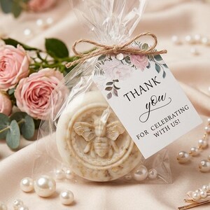 May include: A round, light-colored soap bar with a bee design, packaged in clear cellophane and tied with twine. A tag reads "Thank you for celebrating with us!" Pearls and pink roses surround the soap, creating a romantic theme.