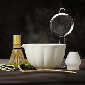 May include: A matcha tea set featuring a white ceramic bowl, a bamboo whisk, a stainless steel sieve, a bamboo scoop, and a white ceramic whisk holder. The set is arranged on a dark wooden surface, with a dark background.